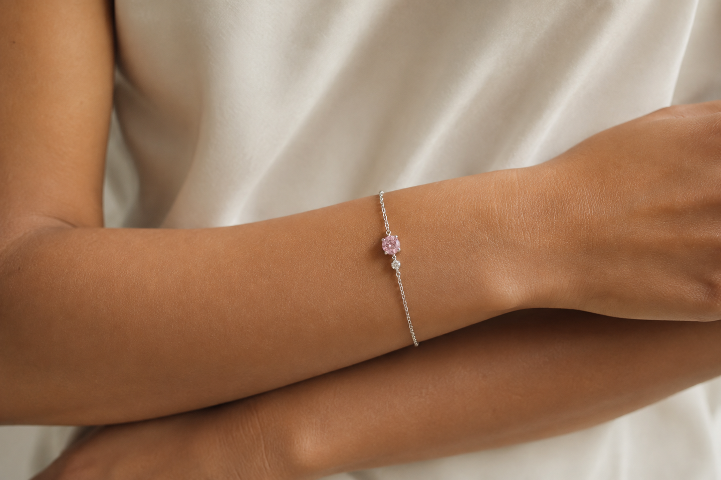 A JOET delicate silver chain bracelet with a small pink charm, worn on a person's wrist against a soft cream-colored fabric background.