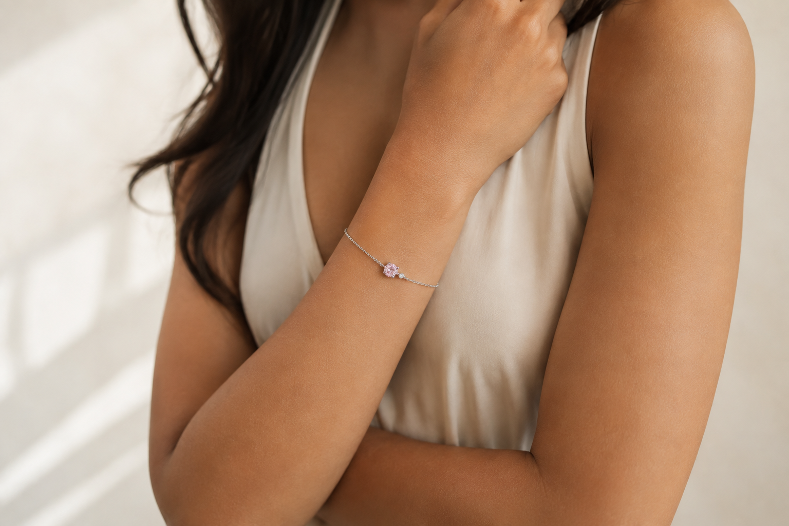 A close-up lifestyle shot of a woman wearing a delicate JOET silver-toned bracelet with a small pink crystal pendant, paired with a cream-colored sleeveless top.